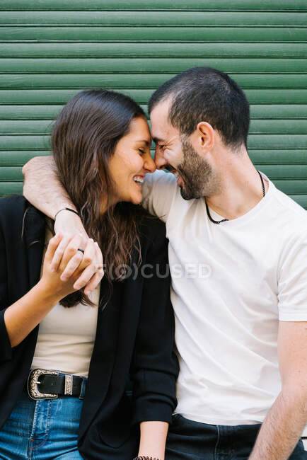 Alegre pareja hispana joven enamorada en ropa casual riendo con los ojos cerrados mientras abraza y toca frentes cerca de la pared verde en la calle de la ciudad - foto de stock