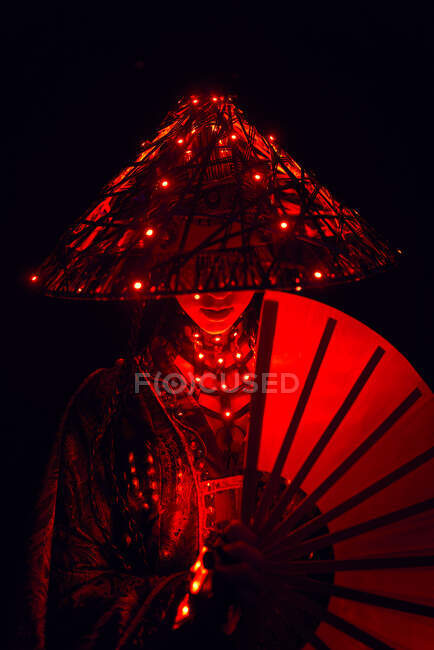 Unerkennbare Frau in authentischem Outfit und traditionellem Hut mit leuchtenden Lampen im dunklen Studio mit Fächer in der Hand auf schwarzem Hintergrund — Stockfoto