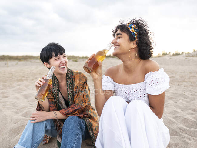 Позитивная многонациональная подруга, пьющая холодное пиво, сидя на песчаном берегу моря и смеясь летним вечером — стоковое фото