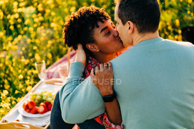 Joven mujer afroamericana con el pelo rizado abrazando y besando novio con los ojos cerrados durante el picnic en floreciente prado en el día soleado - foto de stock