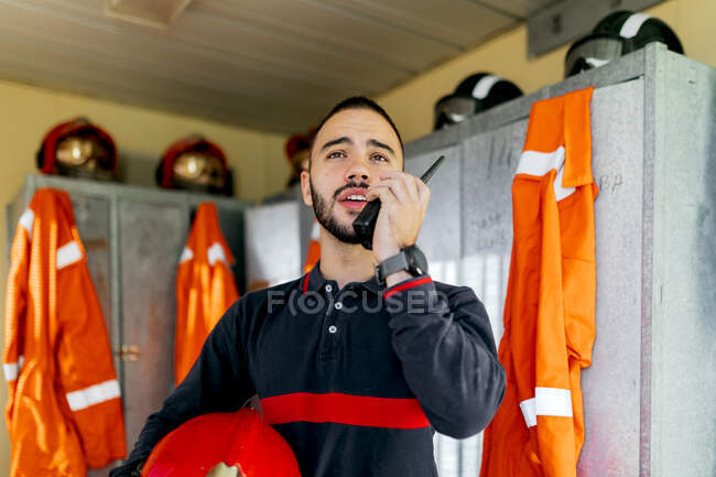 Pompier masculin athlétique ethnique sérieux avec un casque en main debout près de la rangée de casiers en métal avec des casques sur le dessus et un uniforme orange suspendu tout en conversant à l'aide d'un talkie-walkie — Photo de stock