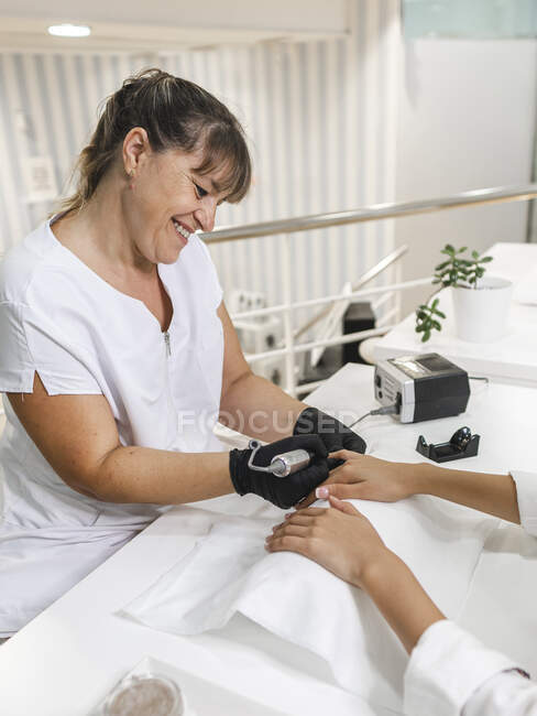 Vista lateral da manicure feminina sorridente em luvas aplicando manicure com ferramentas elétricas em pregos do cliente de conteúdo jovem à mesa — Fotografia de Stock