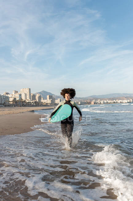 Joven surfista femenina reflexiva en traje de neopreno con tabla de surf corriendo mirando hacia la orilla del mar bañada por el mar ondulante - foto de stock