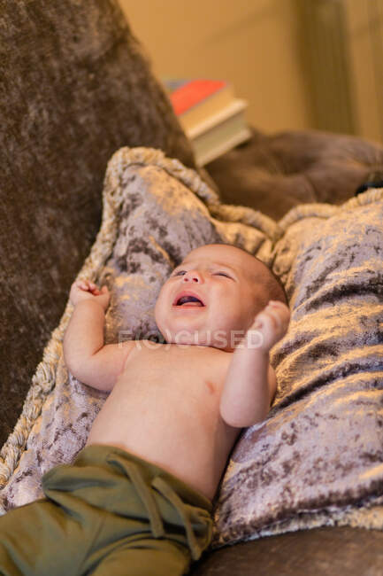 Alto ángulo de adorable bebé sin camisa molesto llorando mientras está acostado en un cojín suave en un cómodo sofá en casa - foto de stock