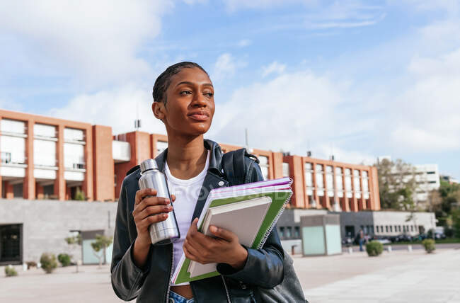 Studentessa afroamericana pensierosa con thermos e un mucchio di libri di testo che distolgono lo sguardo mentre si trova in strada vicino all'edificio universitario — Foto stock