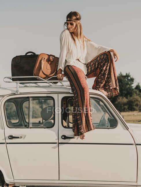 Вид всего тела сбоку стильной женщины-хиппи в одежде, стоящей на белом старом автомобиле с багажником во время поездки на природу — стоковое фото