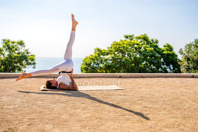 Vista lateral de hembra flexible en ropa deportiva realizando Eka Pada Sarvangasana en estera en suelo seco durante sesión de yoga en parque contra árboles verdes y cielo azul sin nubes a la luz del sol - foto de stock