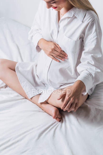 Зверху анонімний молодий вагітна ніжна жінка торкається животик, сидячи на ліжку — стокове фото