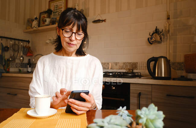 Mujer satisfecha en anteojos mensajes de texto en el teléfono inteligente mientras está sentado en la mesa con una taza de café en la mesa en la cocina - foto de stock