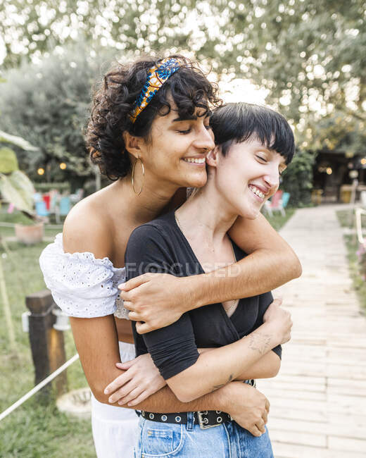 Позитивна любляча багатоетнічна пара гомосексуальних жінок обіймається закритими очима, стоячи на шляху в літньому парку — стокове фото