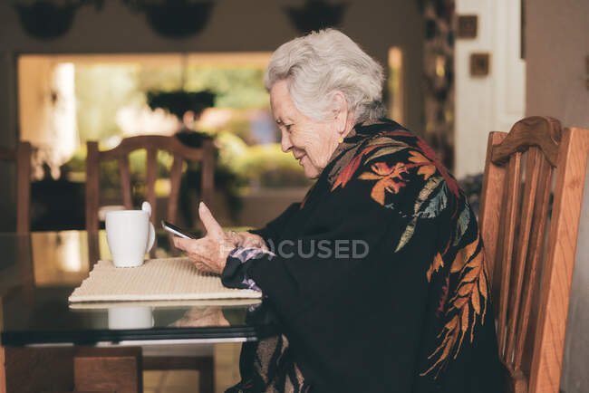 Вид сбоку улыбающейся пожилой женщины в теплой одежде, сидящей за столом с табличкой и чашкой чая, смотрящей на экран — стоковое фото