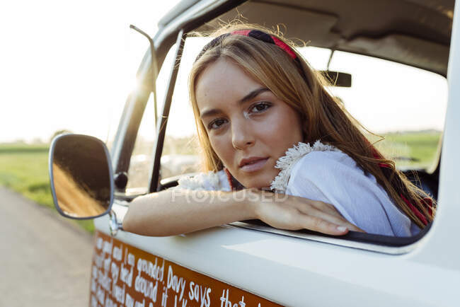 Linda chica rubia sentada dentro de una furgoneta vintage y apoyada en la ventana en un día soleado - foto de stock