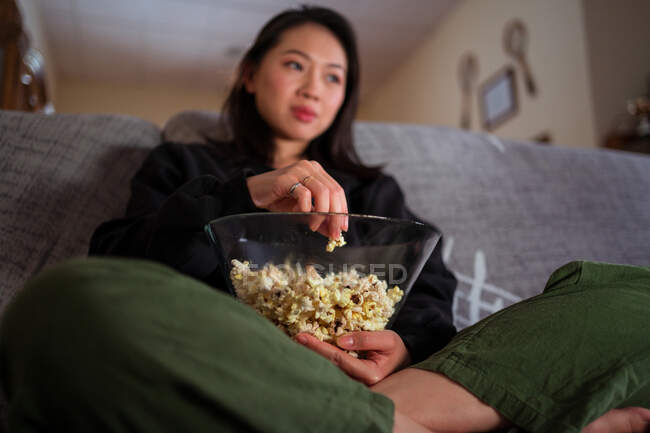Junge Asiatin in lässiger Kleidung sitzt mit überkreuzten Beinen auf der Couch und isst zu Hause Popcorn — Stockfoto