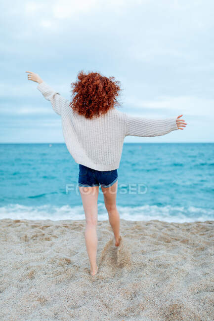Vista posteriore piena lunghezza del viaggiatore femminile a piedi nudi irriconoscibile in piedi sulla costa sabbiosa bagnata da onde schiumose di mare blu — Foto stock