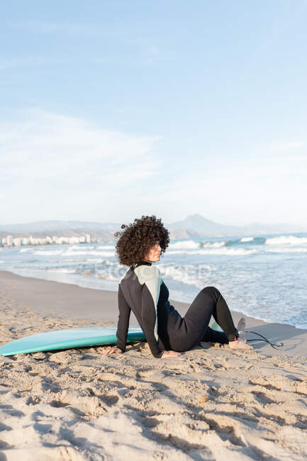 Вид збоку повне тіло босоніж жіночого серфера в гідрокостюмі, що сидить на піщаному пляжі біля моря, що махає — стокове фото