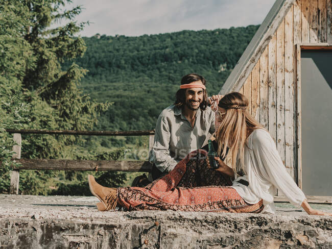 Позитивная пара хиппи в одежде в стиле бохо, сидящая рядом с деревянным строением против леса с деревьями во время путешествия по природе — стоковое фото