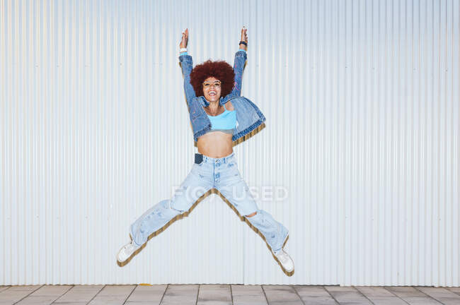 Corpo inteiro de mulher alegre com penteado afro vestindo roupas elegantes pulando com pernas levantadas e braços no fundo branco na rua — Fotografia de Stock