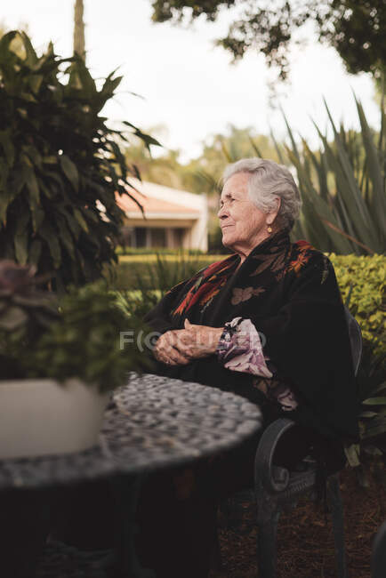 Vista laterale della donna anziana pensierosa con i capelli grigi che avvolgono in plaid seduto al tavolo da giardino e che distoglie lo sguardo nella giornata di sole — Foto stock