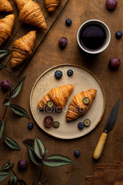 Сверху вкусные свежие круассаны подаются на тарелке с фруктами, помещенными возле чашки чая на деревянном столе в утреннее время в светлом помещении — стоковое фото