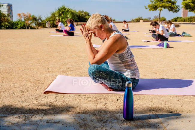 Side view of calm woman with short hair in activewear doing Padmasana with Namaste hands while sitting on yoga mat during outdoors practice in sunny day — Stock Photo