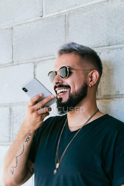 Счастливый бородатый парень с татуировками в черной футболке и солнцезащитными очками, стоящий у стены здания и записывающий аудиосообщение на смартфон при дневном свете — стоковое фото