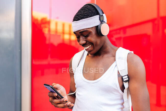Homem afro-americano feliz em roupas brancas ouvindo música em fones de ouvido sem fio e mensagens no telefone celular, enquanto em pé contra a parede vermelha na cidade — Fotografia de Stock