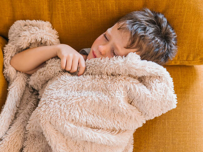Vista superior del niño enfermo con gripe acostado con los ojos cerrados debajo de la manta en el sofá y durmiendo en la sala de estar en casa - foto de stock