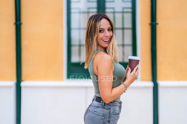 Positive female with paper cup of takeaway coffee smiling and looking at camera while standing near building on street of city — Stock Photo