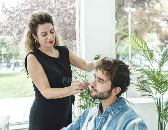 Content female master in black clothes cutting male client in barbershop against window — Stock Photo