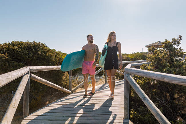 Corpo inteiro de casal esportivo com pranchas de surf passeando juntas no caminho de madeira perto de plantas verdes antes do treinamento em resort tropical — Fotografia de Stock