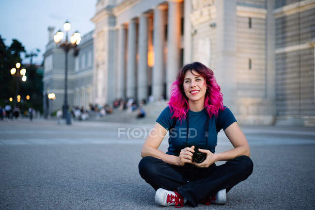 Ganzkörper-Fotografin mit rosa Haaren und Fotokamera in der Hand, während sie auf dem Gehweg in der Nähe eines alten Gebäudes in der Stadt sitzt — Stockfoto