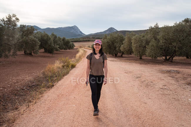 Vista frontal de cuerpo completo de viajera asiática caminando mientras está de pie en la plantación con exuberantes olivos verdes - foto de stock