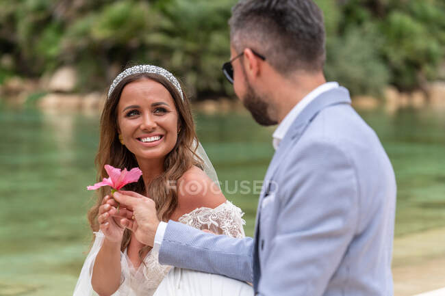 Позитивная супружеская пара в свадебном наряде и солнцезащитных очках, сидящая на каменной лестнице возле озера и зеленых пальм и растений, глядя друг на друга и дарящих цветок — стоковое фото