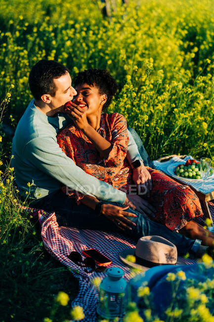 Liebendes multiethnisches Paar, das sich beim Picknick an einem sonnigen Sommertag auf einer Wiese mit Blumen anschaut — Stockfoto