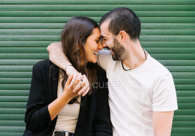 Fröhlich verliebtes junges hispanisches Paar in lässiger Kleidung lacht mit geschlossenen Augen, während es sich in der Nähe der grünen Mauer auf der Stadtstraße umarmt und die Stirn berührt — Stockfoto