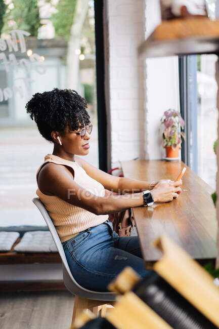 Vista lateral da confiante jovem afro-americana millennial feminino com cabelos encaracolados escuros em roupas da moda e óculos de sol sentados à mesa no café moderno e mensagens no smartphone — Fotografia de Stock