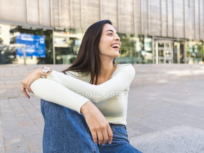 Happy woman with brown hair in casual clothes sitting on concrete border and embracing knee on city street in daylight — Stock Photo