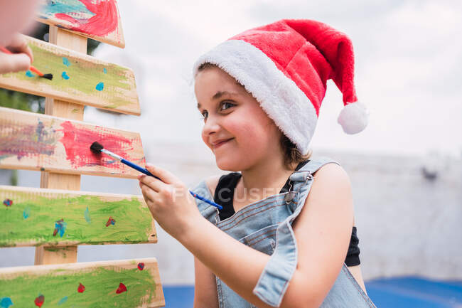 Fille positive en rouge Santa chapeau peinture avec pinceau sur arbre de Noël décoratif coloré pendant la préparation des vacances dans la salle de lumière — Photo de stock