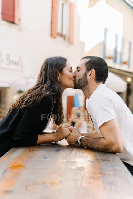 Вид сбоку на влюбленную молодую этническую пару в повседневной одежде, держащуюся за руки и целующуюся, сидящую на городской улице — стоковое фото