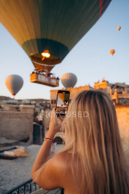 Vue arrière d'une touriste féminine méconnaissable debout à la clôture et prenant des photos de montgolfières sur smartphone — Photo de stock