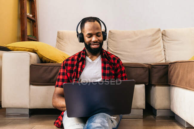From below ethnic male with beard in wireless headphones surfing internet on netbook while sitting on the floor of the house — Stock Photo