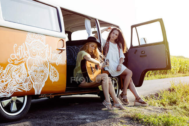 Dos chicas guapas vestidas con ropa de verano divirtiéndose fuera de una furgoneta y tocando la guitarra - foto de stock