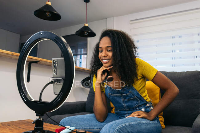 Sorridente femmina nera sul divano durante l'utilizzo di smartphone su lampada ad anello a LED vicino a luci professionali su treppiedi — Foto stock