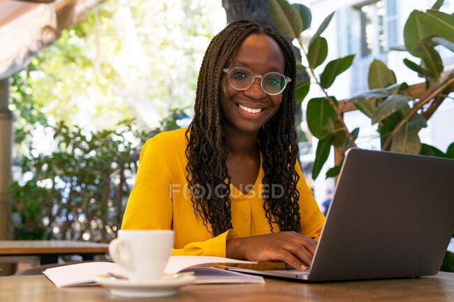 Positif Afro-Américaine pigiste naviguant sur Internet sur netbook tout en étant assis à table avec boisson et ordinateur portable dans la cafétéria extérieure — Photo de stock