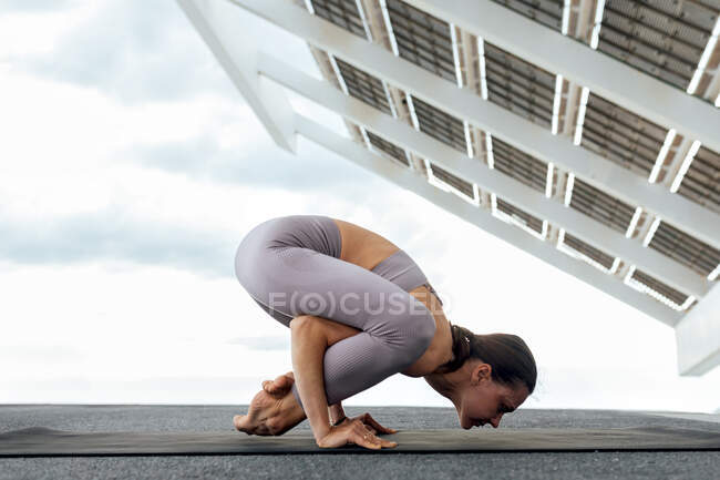 Full body side view of flexible female in sportswoman performing Bhujapidasana posture on street near solar panel during yoga in city — Stock Photo