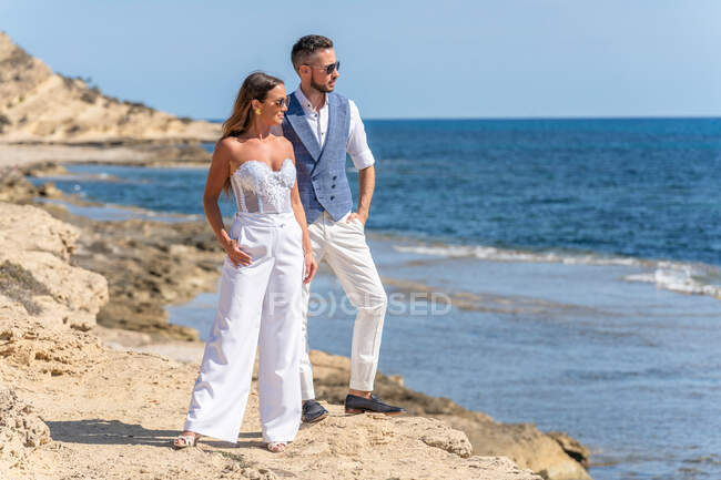 Full body of young couple in elegant outfit and sunglasses standing near sea on hill on rocky coast and looking away under blue sky in sunny day — Stock Photo