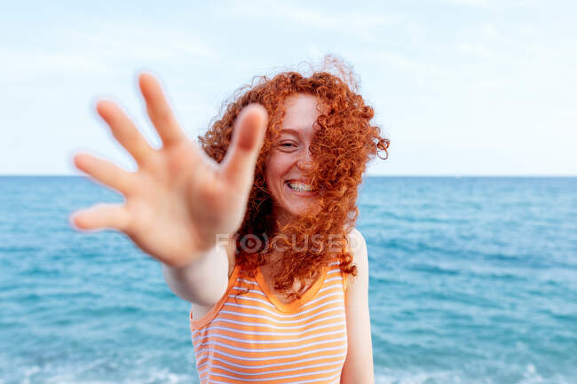 Оптимістична молода жінка з літаючим імбирним волоссям досягає руки до камери на узбережжі блакитного розривного моря — стокове фото