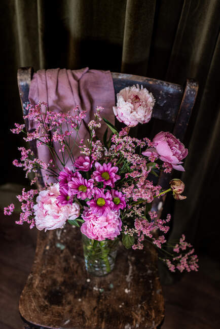 Strauß frischer bunter Pfingstrosen und Chrysanthemen in Glasvase auf verwittertem Holzstuhl in der Nähe von Vorhängen im hellen Raum — Stockfoto