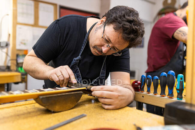 Professional master in apron standing near table with instruments and tools while tuning electric guitar in modern garage — Stock Photo