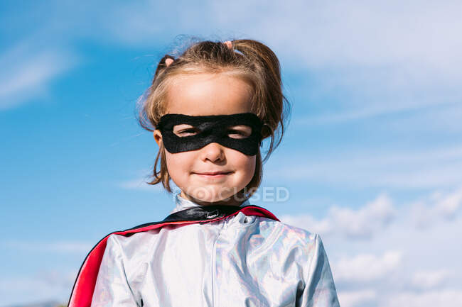 Enfant mignon portant costume de super-héros masqué et masque pour les yeux debout contre le ciel bleu et regardant la caméra — Photo de stock